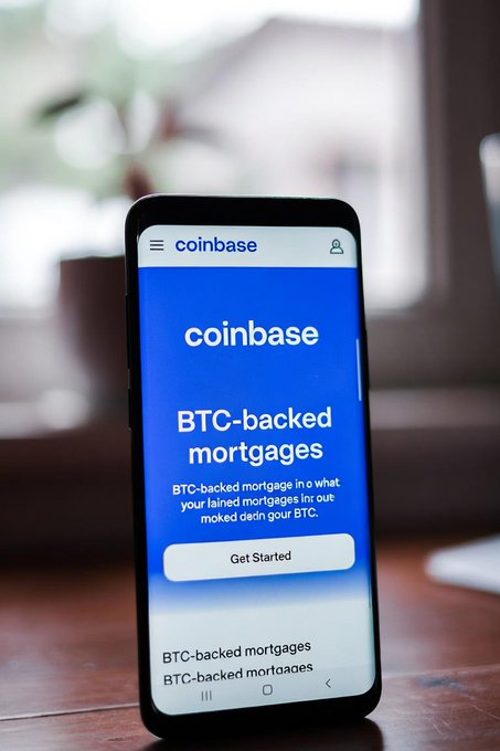 The image shows a smartphone displaying the Coinbase website, which offers Bitcoin-backed mortgages as an alternative way of securing home loans without selling one's Bitcoin. The phone is placed on top of a wooden table or desk.