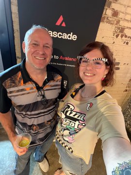 In this image, a man and woman are standing together with their arms around each other, smiling for the camera. They appear to be posing for a photo or attending an event related to Bitcoin Pizza Day. The background of the image features a brick wall, which adds to the overall atmosphere of the scene. There is also a cup placed on a surface in front of them.