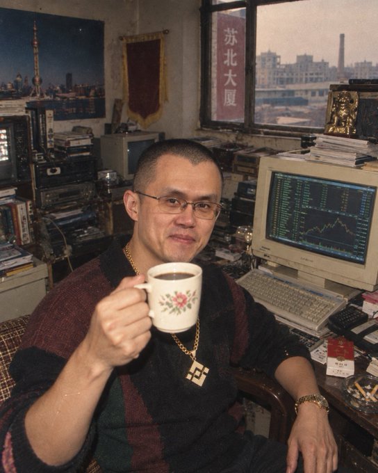 A man wearing glasses and a striped sweater sits at a desk holding a floral mug, with a computer displaying stock charts and Chinese text '苏北大厦' on the window behind him.