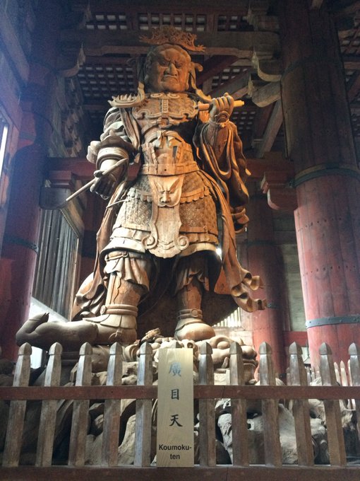 A wooden statue of the Buddhist guardian deity Koumokuten stands inside a temple with red pillars, holding a sword and staff, with a sign reading 'Koumokuten' in front.
