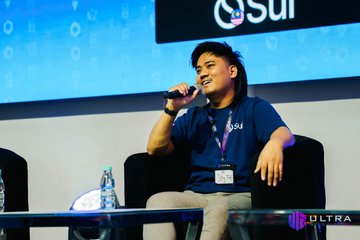 A man in a blue Sui-branded t-shirt speaking into a microphone while seated on stage at what appears to be a conference or panel event.
