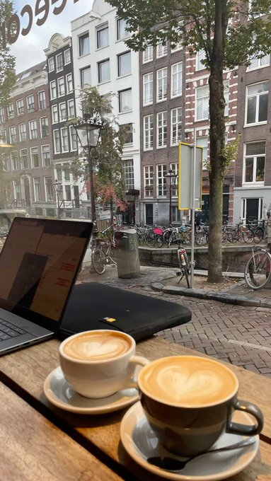 Two cappuccinos with heart-shaped latte art sit on a wooden café table beside an open laptop, overlooking a classic Amsterdam canal scene with bicycles, canal houses, and a lamppost.

The post caption "GM from Amsterdam. Canal views, caffeine, and a fresh day to lock back in." aligns perfectly with the image—the coffee and laptop capture the "caffeine" and "lock back in" work-from-café vibe, while the iconic Dutch canal houses and bikes deliver the quintessential "Amsterdam" canal views mentioned.