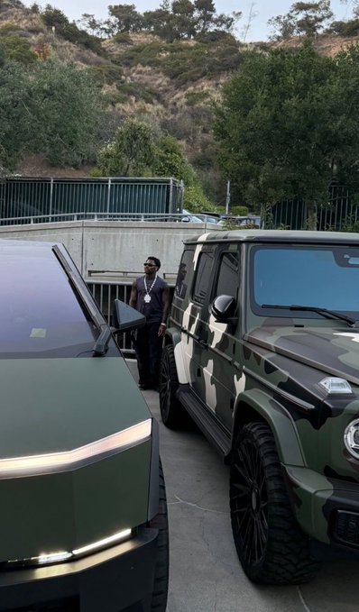 A man in a dark t-shirt stands between two large vehicles outdoors, with a green military-style SUV parked on the right.