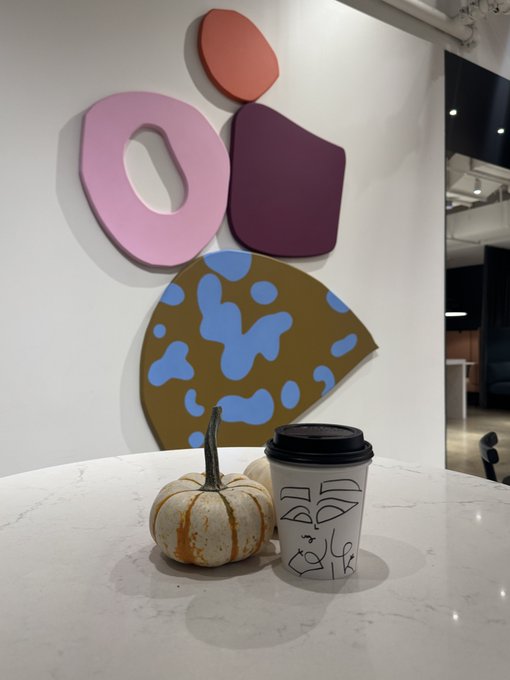 A small pumpkin sits on a white marble table next to a white coffee cup with a black lid and black line-art face design, with abstract colorful wall art hanging in the background.