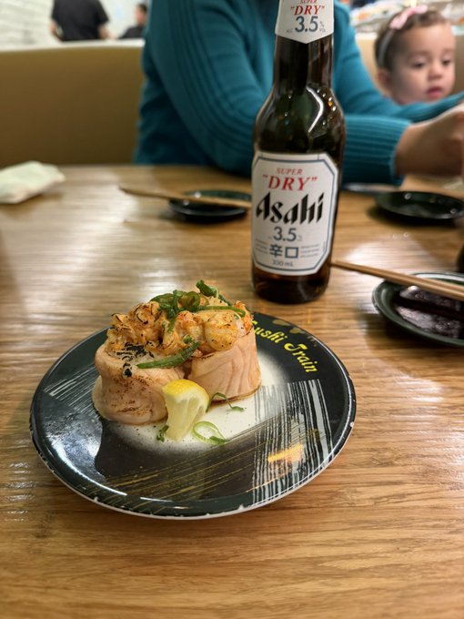 A restaurant table displays a plate of sushi with 'Asahi Chain' printed on it, accompanied by a bottle of Asahi 'Super Dry' 3.5 beer and a child visible in the background.