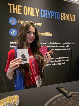 A woman posing at a "THE ONLY CRYPTO BRAND" booth (lhodllife.com), holding hardware wallets in front of a display table with gold coins.