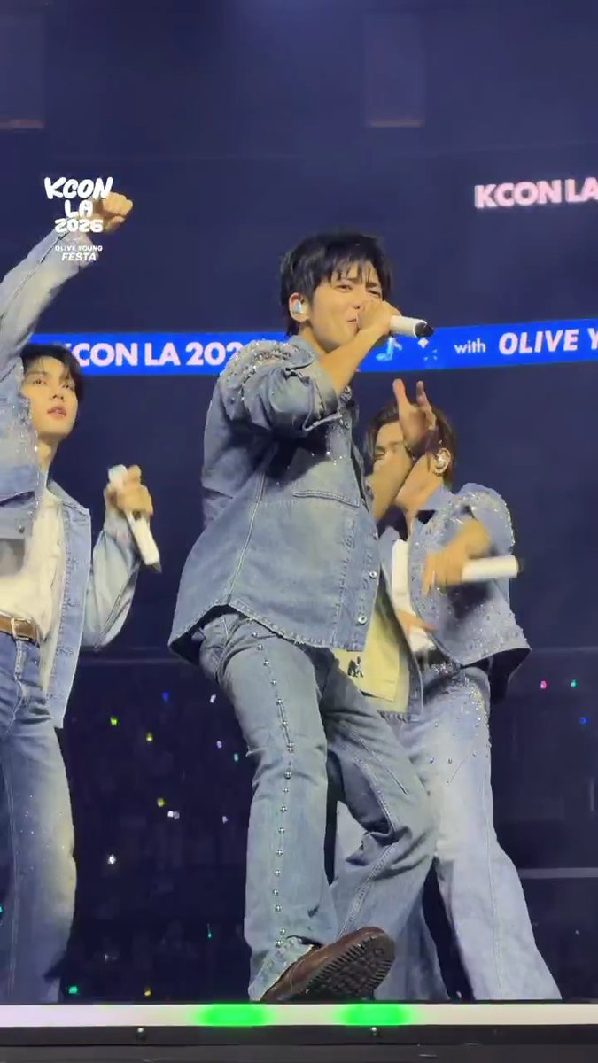 TXT (Tomorrow X Together) performs on stage at KCON LA 2026, with members in coordinating denim outfits singing into microphones in front of a banner displaying "KCON LA 2026" and "with OLIVE." The center member leans forward energetically into the mic, while another member raises a fist in the air, capturing the high energy of the group's live countdown stage performance.