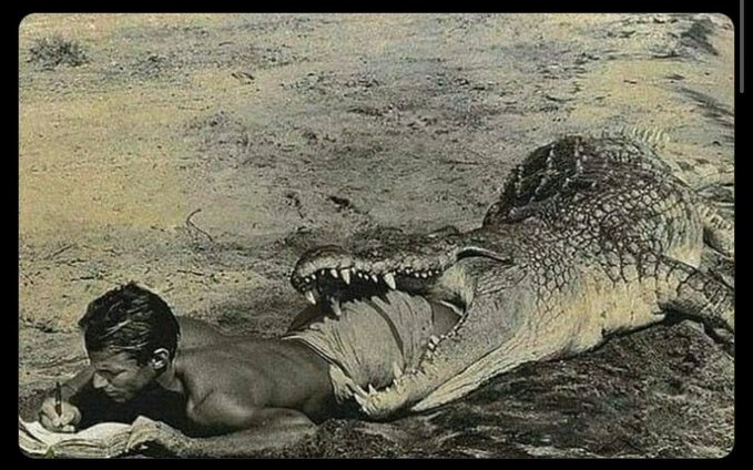 A surreal, edited image showing a shirtless man lying on the ground calmly writing in a notebook while a massive crocodile bites his torso.