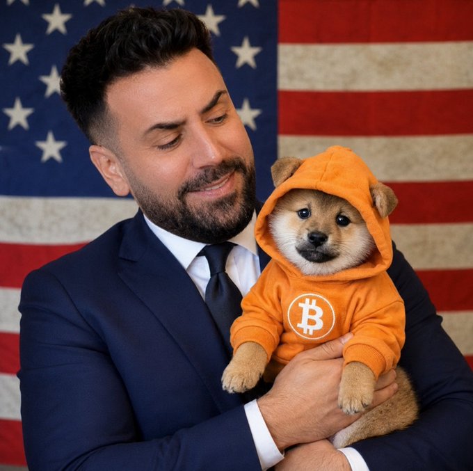 A man in a navy suit and tie holds a Shiba Inu puppy wearing an orange hoodie with a Bitcoin logo, posed in front of an American flag.