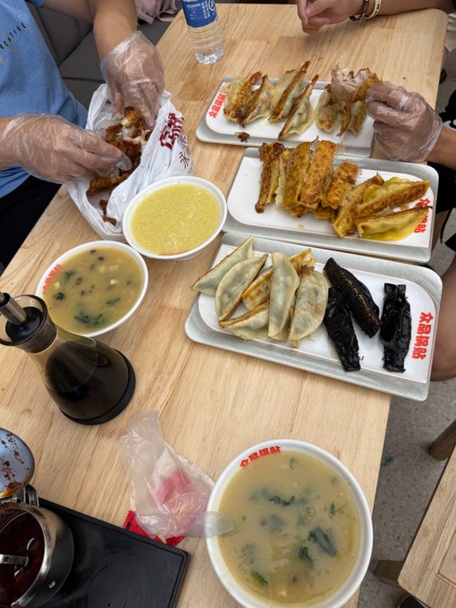 An overhead view of a meal at a wooden table featuring plates of fried and steamed dumplings, bowls of yellow and green soups with greens, and bottles of soy sauce, with two people wearing clear plastic gloves eating.