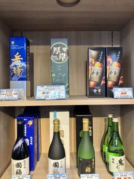 A display shelf in what appears to be a liquor shop or airport duty-free area featuring various premium Japanese sake bottles with elegant packaging and price tags.