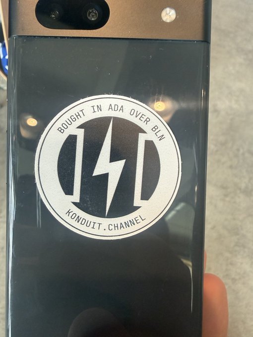 The image shows a person holding a black smartphone with a sticker featuring a lightning bolt symbol, possibly representing Bitcoin Lightning points or ADA over Bluetooth. The phone's case has a logo for Konduit Channel, indicating that the stickers were purchased from them. This demonstrates how blockchain ecosystems can be used to create unique and personalized user experiences by allowing users to customize their devices with custom stickers and logos.