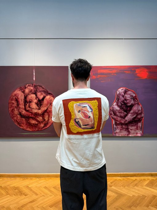 A man wearing a white t-shirt with an artwork design on the back stands viewing two large red-toned paintings in a gallery.
