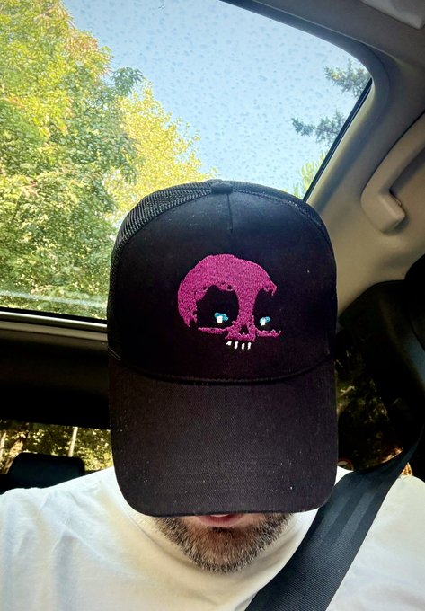 A man wearing a black trucker hat with a pink skull design sits in a car, the hat obscuring his face.