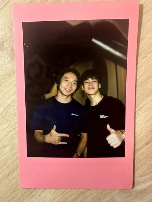 A pink-bordered Polaroid photo of two young men smiling and posing with hand gestures, taken against a graffiti background.