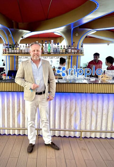 CEO of Flash Payments Nicolas Steiger, a Ripple partner, poses at a Ripple-branded bar during Ripple APAC's client celebration in Singapore, holding a glass of champagne. The "ripple" logo is prominently displayed on the bar, with bottles of spirits lined up behind the counter.