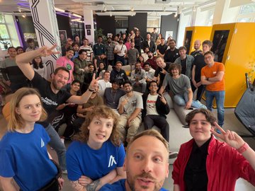The image shows a large group of people posing for a photo, all wearing blue shirts. The group consists of both men and women, with some standing and others sitting down. They are gathered together in an office setting, possibly celebrating or commemorating something. In the background, there is a yellow door that adds to the overall color scheme of the scene.