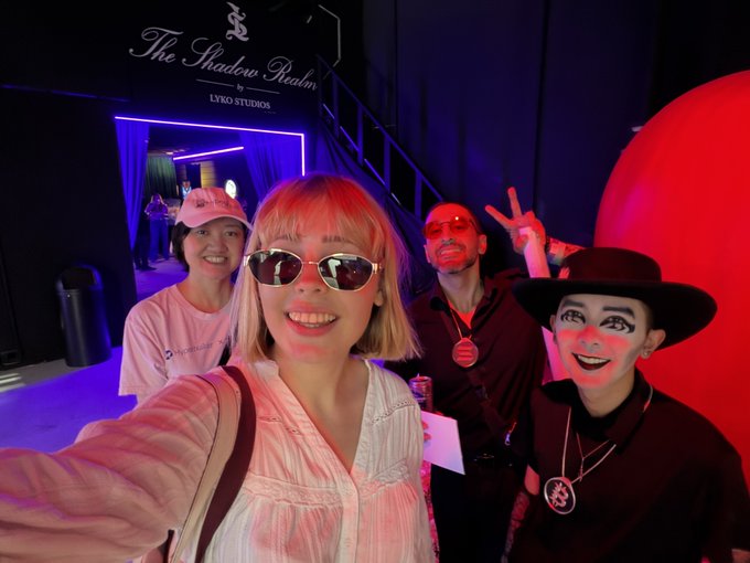 A group selfie taken at 'The Shadow Realm' by LYCO Studios shows several people, including a woman with pink hair and sunglasses, and a performer in a black hat and dramatic makeup.