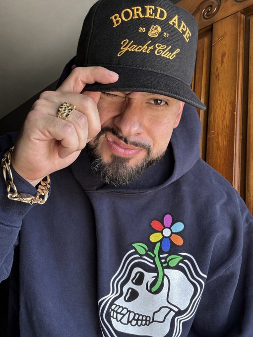 A man wearing a black 'BORED APE 20 21 Yacht Club' cap and a skull-flower hoodie poses with a gold chain and ring visible.
