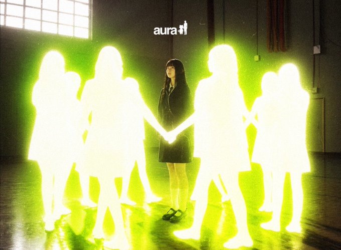 The image shows a dark-haired woman in a school uniform standing at the center of a circle, holding hands with several glowing yellow-green silhouette figures in what appears to be a gymnasium, with the "aura" logo visible at the top—visually referencing the Aura Network community post wishing good morning to its members. The ethereal, luminous figures surrounding her evoke the interconnected, energetic community spirit described in the accompanying "Gm $Aura community" message.