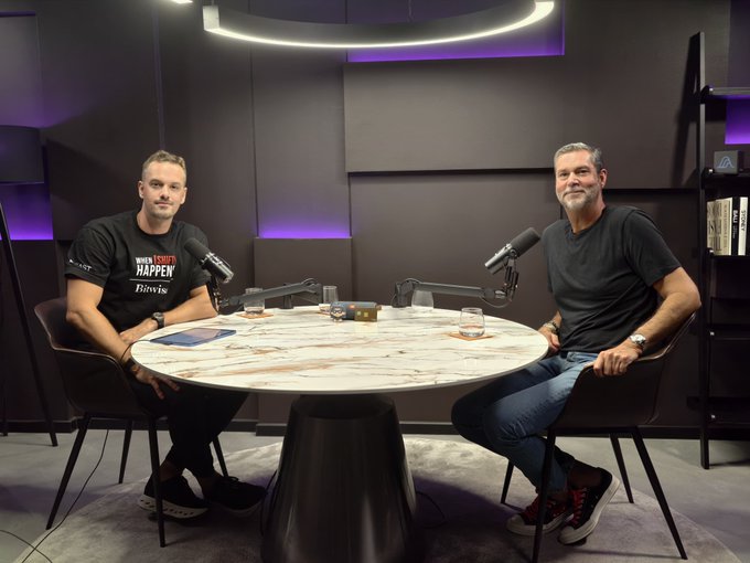 Two men sit across from each other at a marble-topped table in a modern podcast studio, with one wearing a "When Things Happen Bliving" t-shirt. The setup includes professional microphones and a camera between them, suggesting a formal interview or podcast recording session.