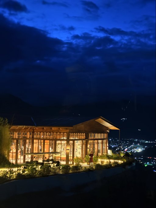A warmly lit, modern glass-walled building perched on a hillside at dusk, overlooking a valley dotted with the distant city lights of a sprawling town below.