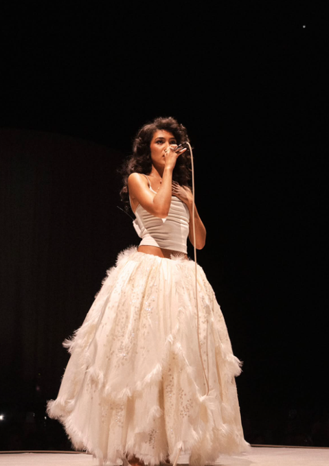 A woman with curly hair, dressed in a white corset top and a flowing lace and feather skirt, performs on stage while singing into a microphone. The image complements the post by visually referencing Olivia Dean, the British singer behind "Man I Need," who is favored to win Record of the Year at the Grammys according to Polymarket traders.