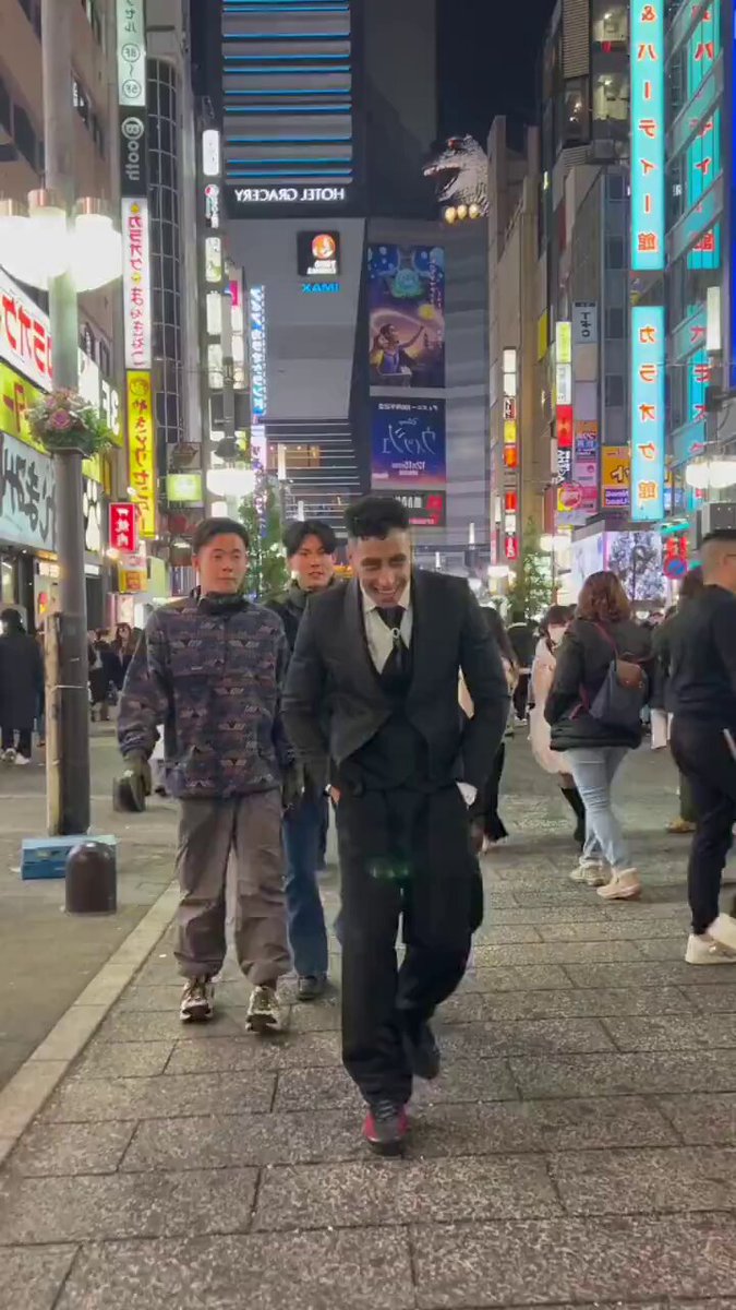 The image shows a man walking down a city street at night, wearing a suit and tie. The surrounding area has many neon signs and buildings lit up with bright lights, creating an urban atmosphere. There are also other people visible in the scene, some of whom appear to be carrying handbags or backpacks. A post on this image reads "$LUNC holders when Terra Luna Classic hits 0.01". This suggests that the photo was taken during a special event or promotion for a product or service related to the movie "Terra Luna Classic", possibly a limited edition release of the film, which is known for its visually stunning scenes and vibrant colors. The neon signs and bright lights in the background further emphasize the connection between the image and the promotional content.