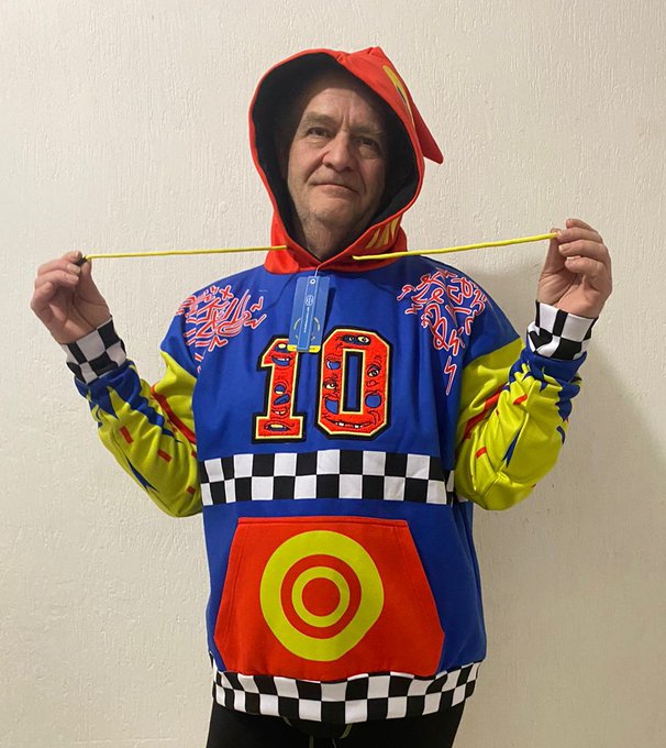 A person wearing a colorful blue and yellow hoodie with the number "10" and a red hood pulls on the drawstrings while posing against a plain wall, looking directly at the camera. The vibrant, cartoon-style clothing visually contrasts with the somber, memorial tone of the post paying tribute to Larry, a "truth teller" who has passed away.