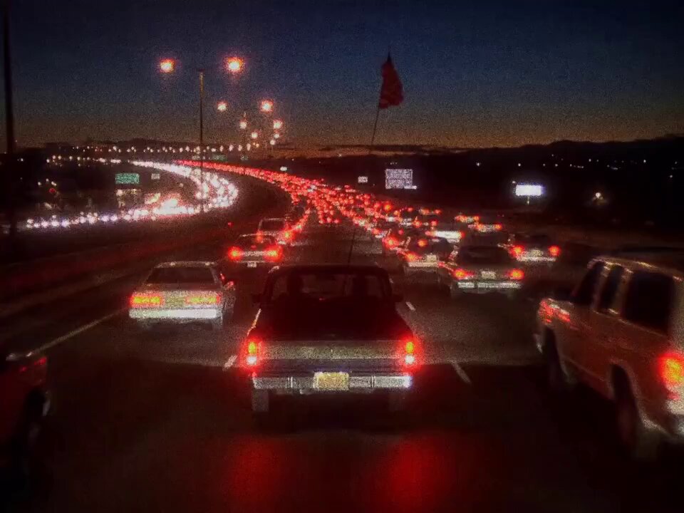 The image shows a busy highway at night with multiple cars driving down the road. The traffic appears to be backed up or stopped due to congestion. There are several cars visible, including one car that has its lights on, indicating that it might be in the middle of an accident or experiencing some other issue. The scene is illuminated by streetlights and car headlights, creating a sense of darkness around the highway.