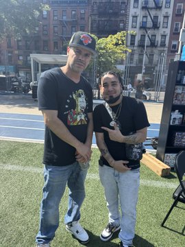 Two men posing together outdoors, one wearing a black t-shirt with colorful graphics and a red cap, the other in a black t-shirt with chains, standing on artificial grass.