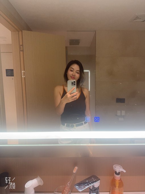 A woman in a dark tank top and light skirt takes a mirror selfie in a modern bathroom, with a smartphone and toiletries visible on the counter below.