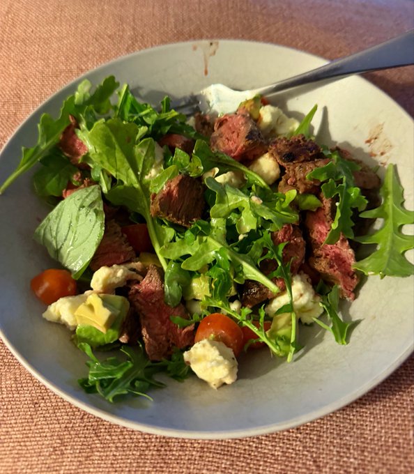 Top-down photo of a hearty salad bowl containing sliced steak, arugula, cherry tomatoes, avocado, and crumbled cheese on a textured pink surface.