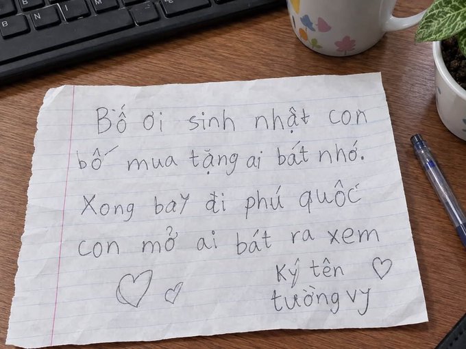 A handwritten note in Vietnamese on lined paper by 'Tường Vy' reads 'Bố ơi sinh nhật con bố mua tặng ai bất nhớ. Xong bay đi phú quốc con mờ ai bắt ra xem' (Dear dad, on my birthday, buy a gift for me. Then fly to Phu Quoc, I'll take you to see), with a keyboard and coffee mug nearby.