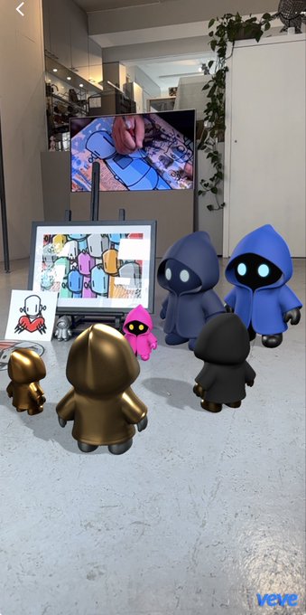 Several small hooded figurines in gold, blue, black, and pink displayed on a counter alongside framed artwork in what appears to be a gallery or shop.