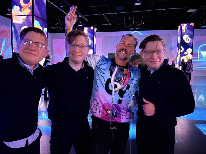The image shows four men posing together, with one man wearing a colorful jacket that has the letters "G" and "M" printed on it. The other three men are standing close by, smiling at the camera. They appear to be enjoying themselves in front of a large screen or projection area, possibly at an event like ApeFest.