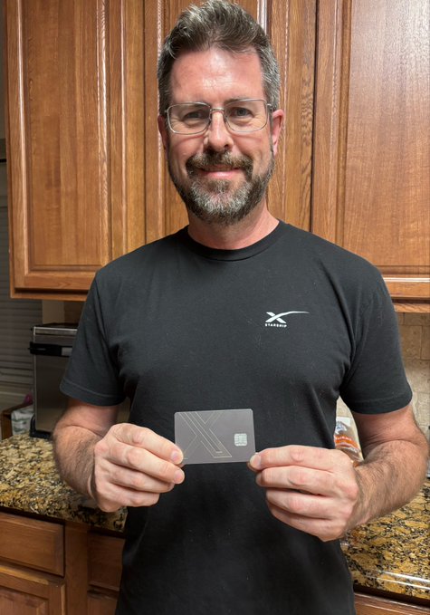 A bearded man wearing glasses and a black t-shirt with a logo holds up an 'X' branded card in front of wooden kitchen cabinets.