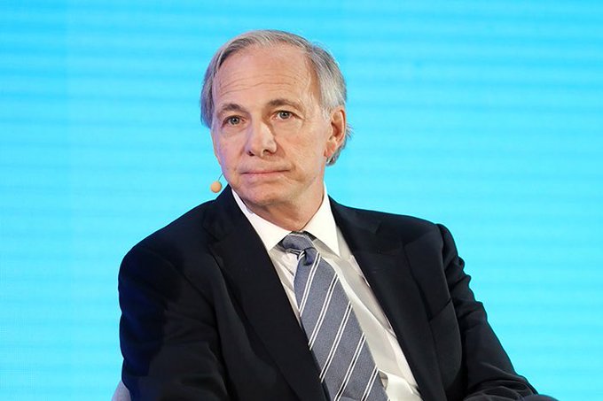 Ray Dalio, the billionaire hedge fund manager, is shown speaking at what appears to be a formal conference or panel discussion, wearing a dark suit with a striped blue tie against a bright blue backdrop. The image visually anchors the accompanying post about Dalio reportedly shifting his investment strategy away from debt-based assets toward hard money like gold and Bitcoin.