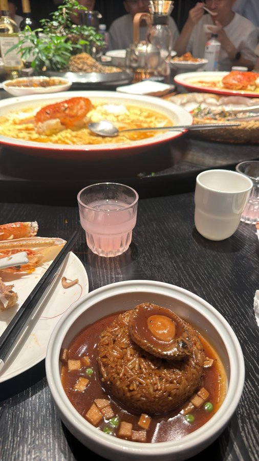 A dinner table spread featuring a bowl of abalone rice with red sauce in the foreground, along with other dishes, crab, and drinks in a restaurant setting.