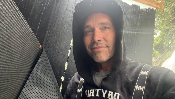 A man wearing a black hoodie with the hood up and a 'Helly Hansen' branded strap takes a selfie, looking toward the camera with a slight smile against a dark, textured background.