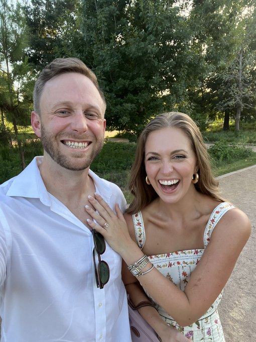 A joyful couple beams with excitement outdoors—likely celebrating an engagement, as suggested by the ring visible on her finger and the "she was in fact a keeper" caption. The man wears a crisp white button-down, while the woman in a floral sundress laughs radiantly, her hand resting on his chest.