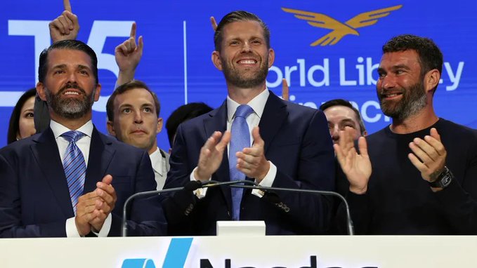 Donald Trump Jr. and Eric Trump smile and applaud at a "World Liberty" financial event, with the company's logo displayed on screen behind them, as their firm faces scrutiny over a new partnership with WorldClaw involving AI models from Chinese companies flagged for national security concerns.