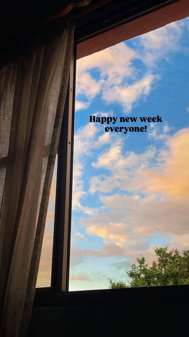A view through a curtained window of a partly cloudy sky at sunset with the overlaid text 'Happy new week everyone!'