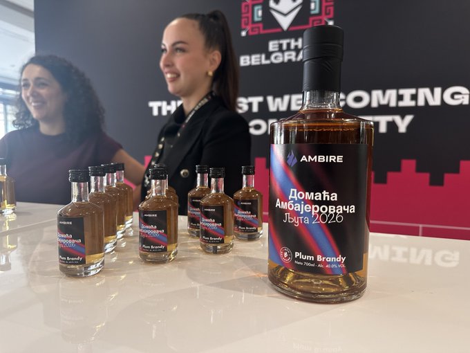 A bottle of AMBIRE 'Домашня Амбалеровча Љута 2026' Plum Brandy sits on a white table with smaller sample bottles in front, while two smiling women stand in the background near a banner for 'ETH Belgr... The Fastest Web Coming...'.