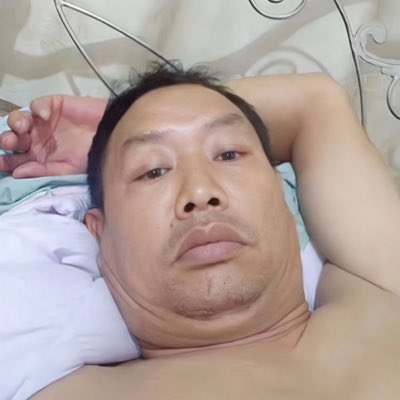 A shirtless man lies on a bed, looking at the camera with a neutral expression, his arm resting behind his head.