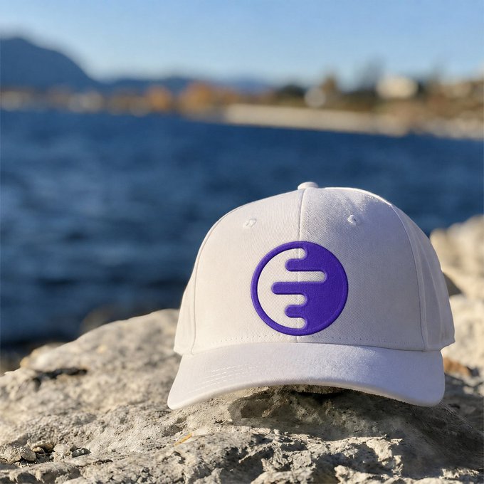 A white baseball cap with a purple circular logo featuring three horizontal bars sits on a rock with a lake and mountains blurred in the background.