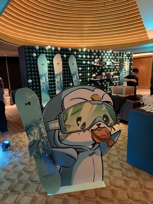 A display at an event features a cartoon sloth character wearing ski goggles alongside a snowboard with a 'Hypoallergenic' label, set against a backdrop decorated with plush toys.
