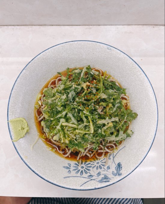 A top-down view of a bowl of noodles topped with fried greens, served in a dark broth with a side of wasabi, presented in a white ceramic bowl with blue floral patterns.