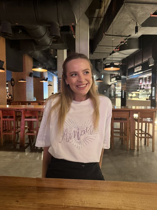 A smiling young woman poses in a modern food hall or bar setting, wearing a white t-shirt featuring "Auburn" in stylized lettering with a sunburst design, while the post playfully compares her to a Doodles NFT character toy.