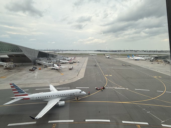 An American Eagle regional jet taxis on an airport tarmac with other aircraft visible at gates in the background under a cloudy sky.
